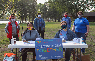 Photo of people staffing the Welcoming Week table. A sign in front of the table says "Creating Home Together."