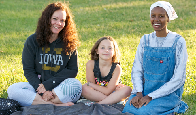 A woman with her young daughter--who are white--sit on a blanket in a park with a young woman who is black.