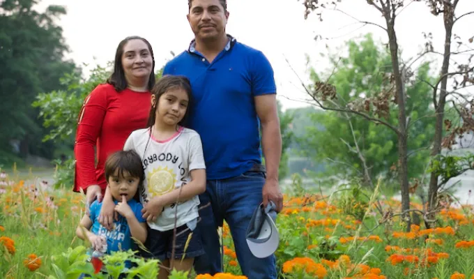 Photo of a Latino Minnesotan family: dad, mom, an elementary-school daughter and a pre-school son. There are orange flowers, trees, and a river in the background.