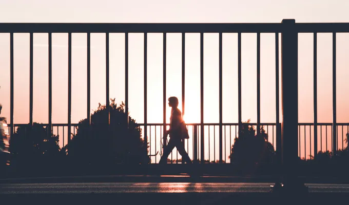Photo of a man in silhouette walking on a sidewalk against a fence, with a sunset in the background. Photo by Kasper Rasmussen on upsplash.