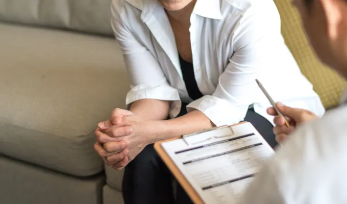 Cropped photo of an adult sitting on a couch, speaking to another adult with a clipboard