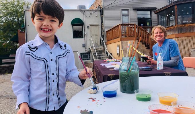 Photo of a young boy smiling at the camera. He is participating in an art project. A woman who is helping at the table smiles in the background