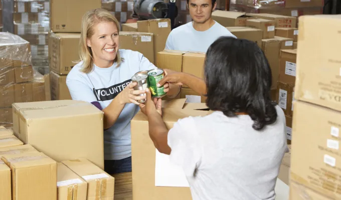 Three adult volunteers, standing amongst cardboard boxes and handing canned food to one another