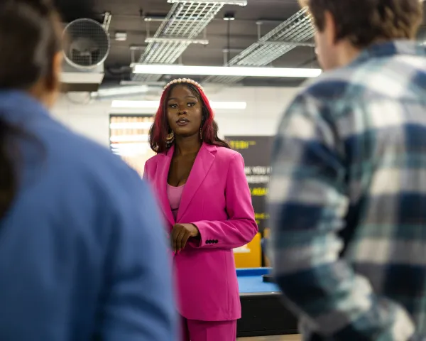 A young Black woman in a bright pink pantsuit in the background, facing the camera, addresses two colleagues in the foreground with their backs to the camera.