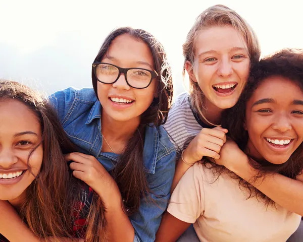 Photo of four teenage girls around 14. They have their arms around each other and are smiling at the camera.