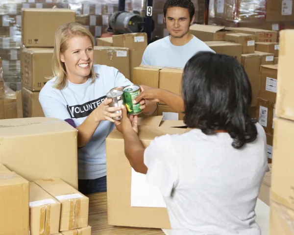 Three adult volunteers standing amongst boxes, handing canned food to each other