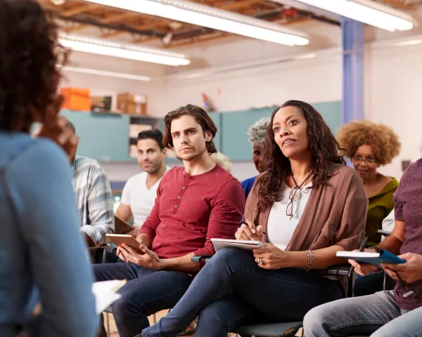 Photo of adults sitting in classroom desks, focused on a speaker at the front of the room