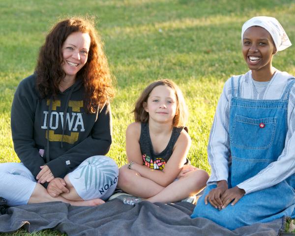 Two smiling women--one white, one black, sit on a blanket in a park. A young white girl sits between them