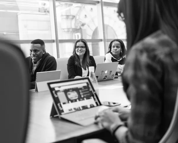Black and white photo of adults sitting around a workplace meeting table, with laptops in front of them