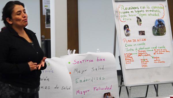 Hispanic woman facilitating a meeting. Items on the flipchart next to her are in Spanish.