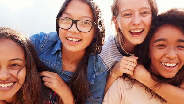 Photo of four teenage girls around 14. They have their arms around each other and are smiling at the camera.