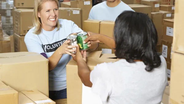 Three adult volunteers standing amongst boxes, handing canned food to each other